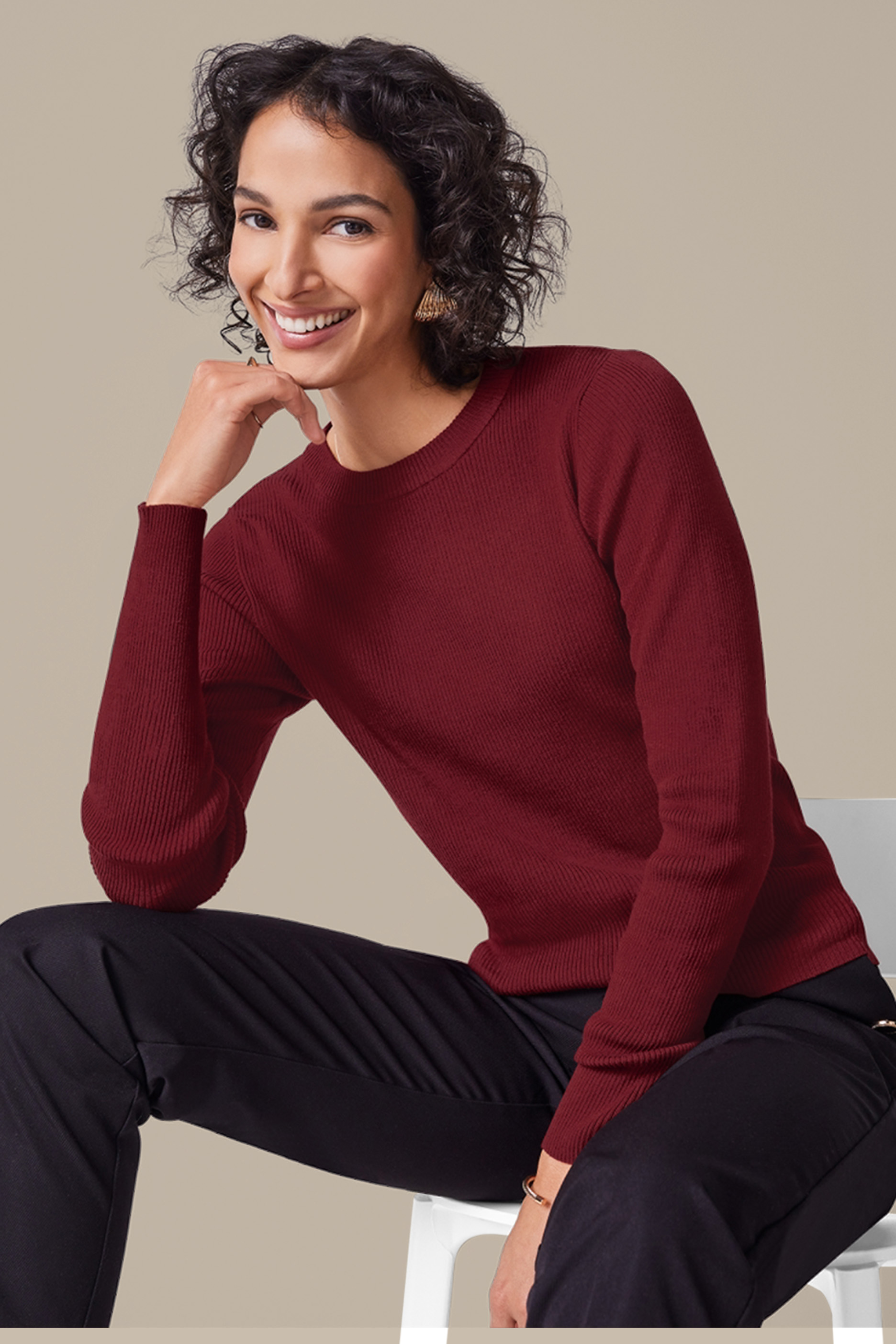 Sieviete sarkanā rievotā džemperī un melnās biksēs sēž un smaida.