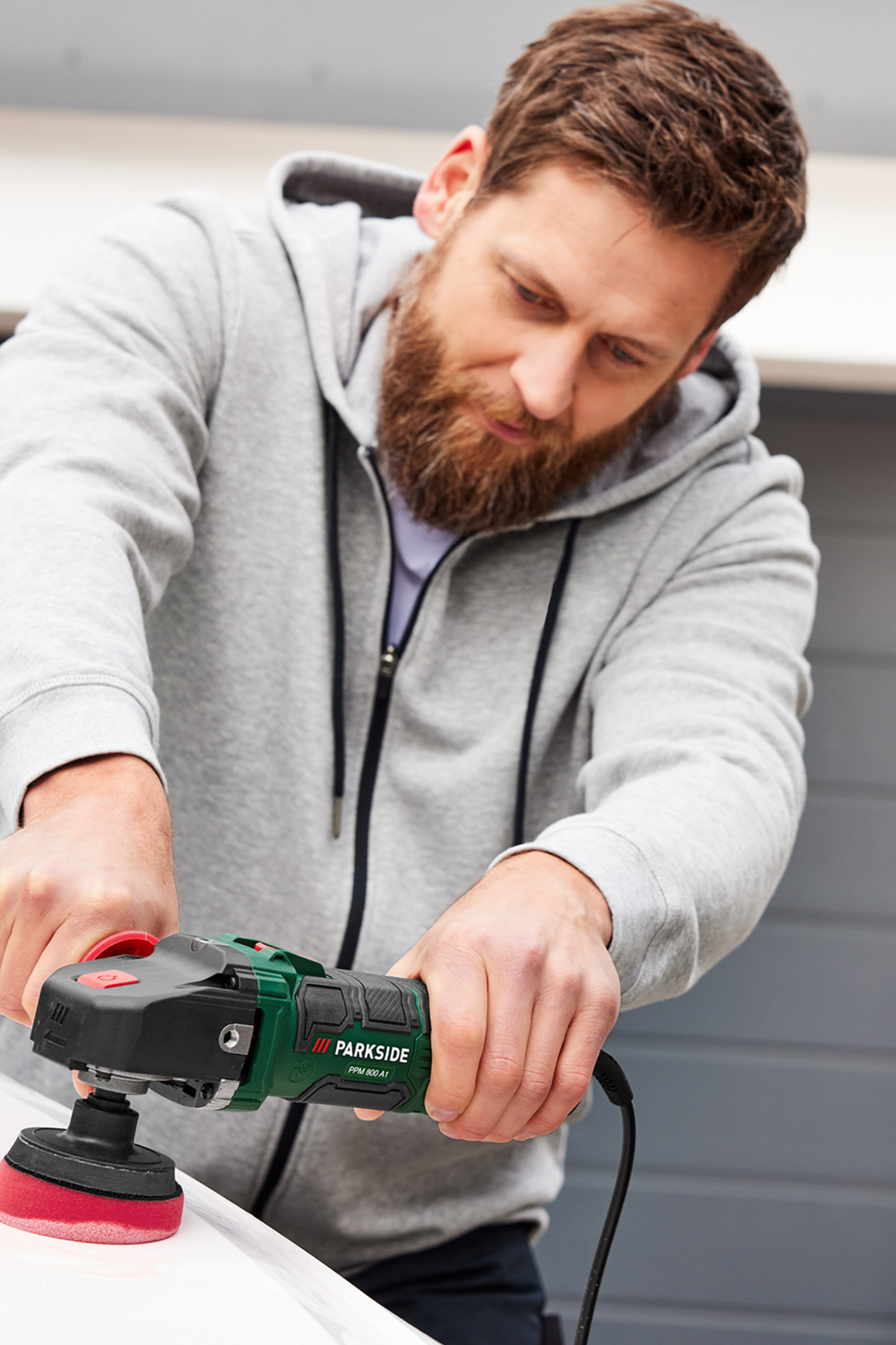 Vīrietis pelēkā džemperī pulē virsmu ar Parkside pulēšanas mašīnu.