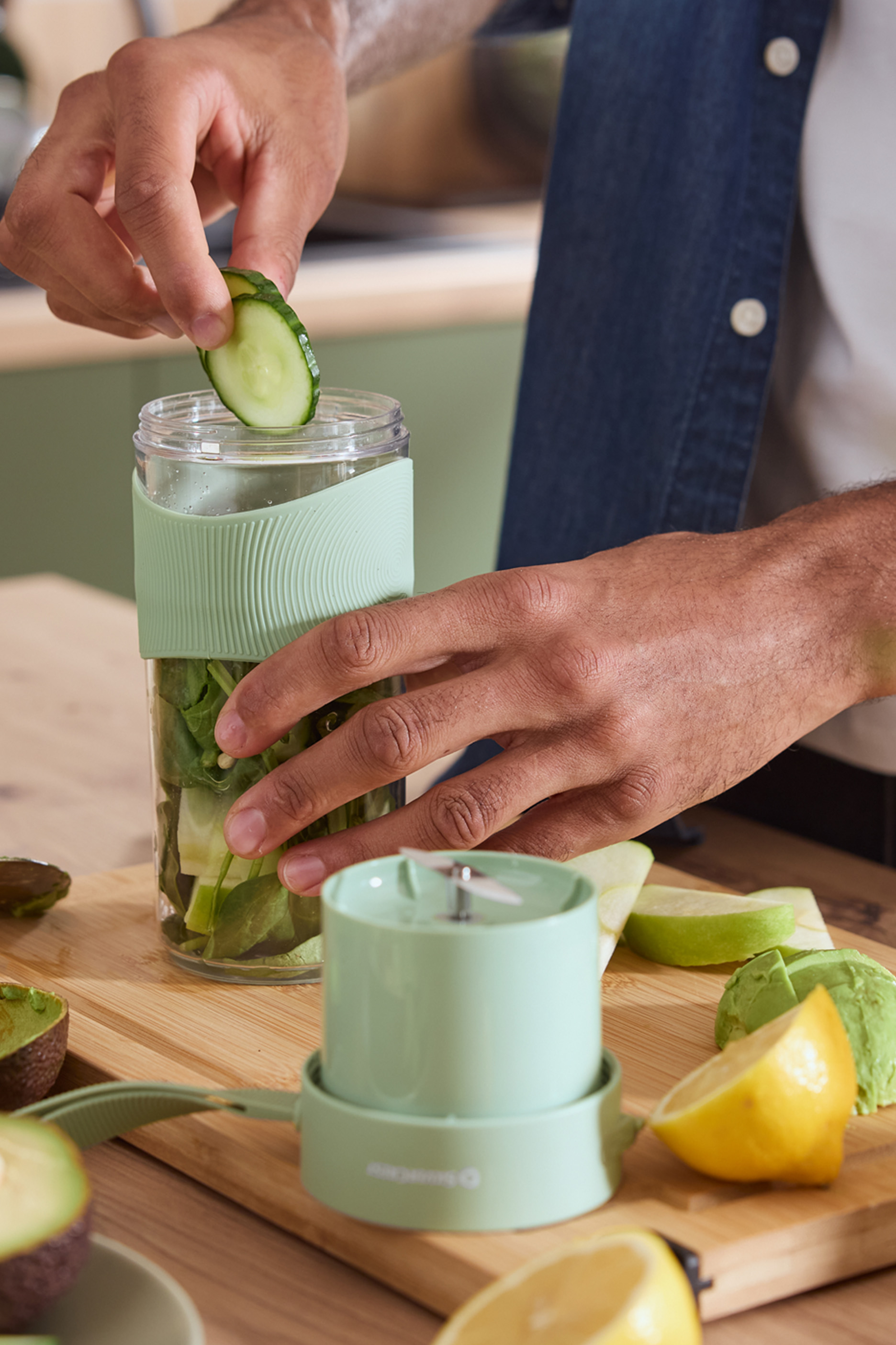 Cilvēks pievieno gurķi pārnēsājamam blenderim ar zaļumiem, blakus augļi.