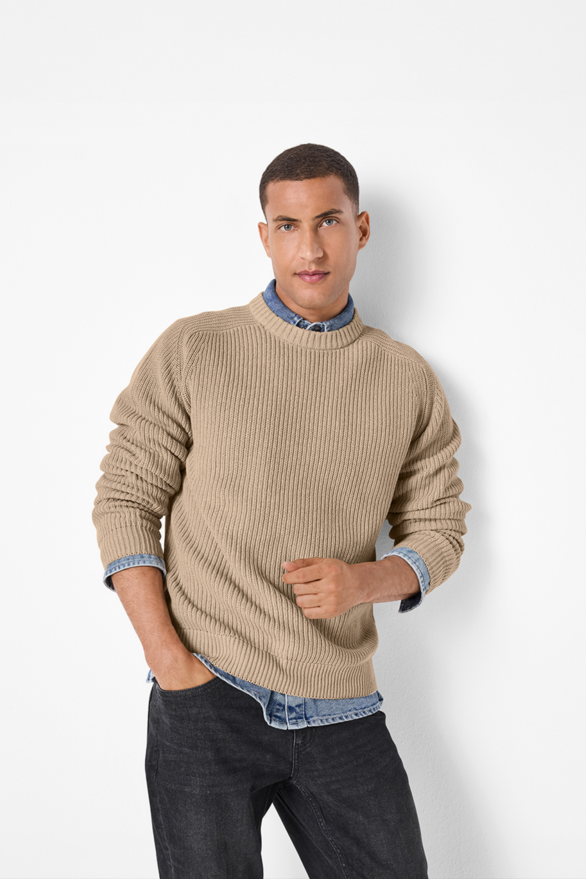 Vīrietis bešā adītā džemperī un tumšās džinsās, ar roku kabatā.