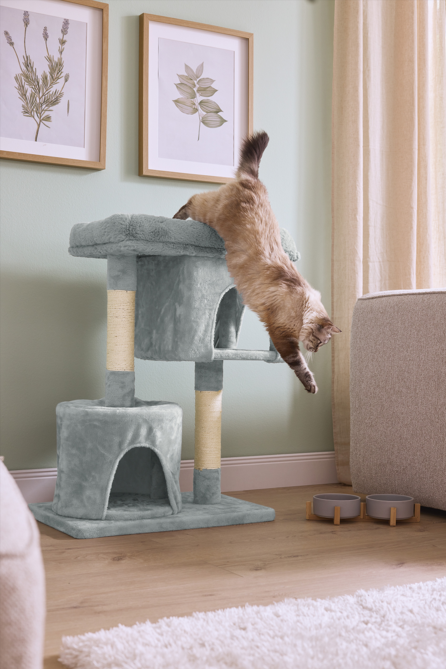 Kaķis lec no pelēkas kaķu mājas ar skrāpējamiem stabiem, blakus bļodām un paklājam.