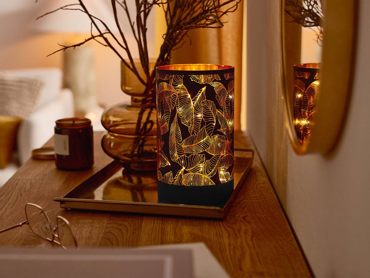 Декоративный светодиодный фонарь с узором из листьев, ваза с ветками и свеча на деревянном столе.