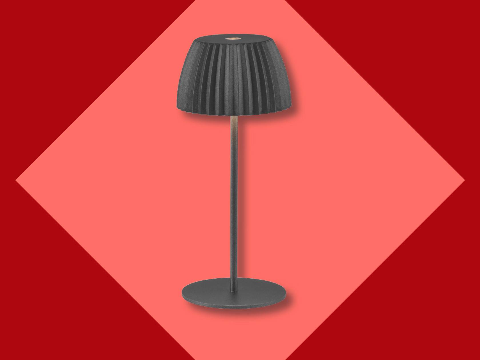 Galda lampa ar melnu krokotu abažūru un tievu kāju uz sarkana fona.