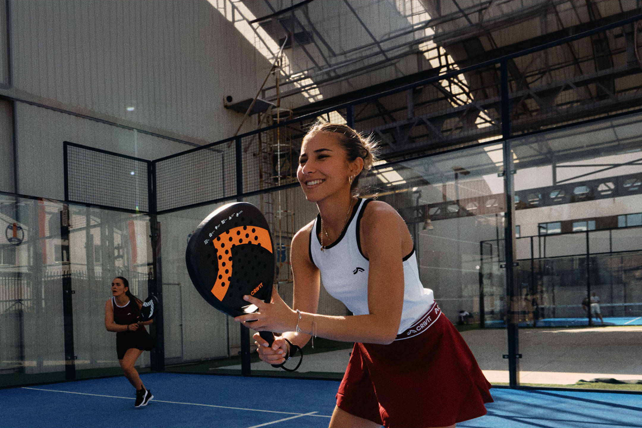 MI ģenerēts: Divas sievietes spēlē padel tenisu, viena smaida un tur raketi, uz zila laukuma.