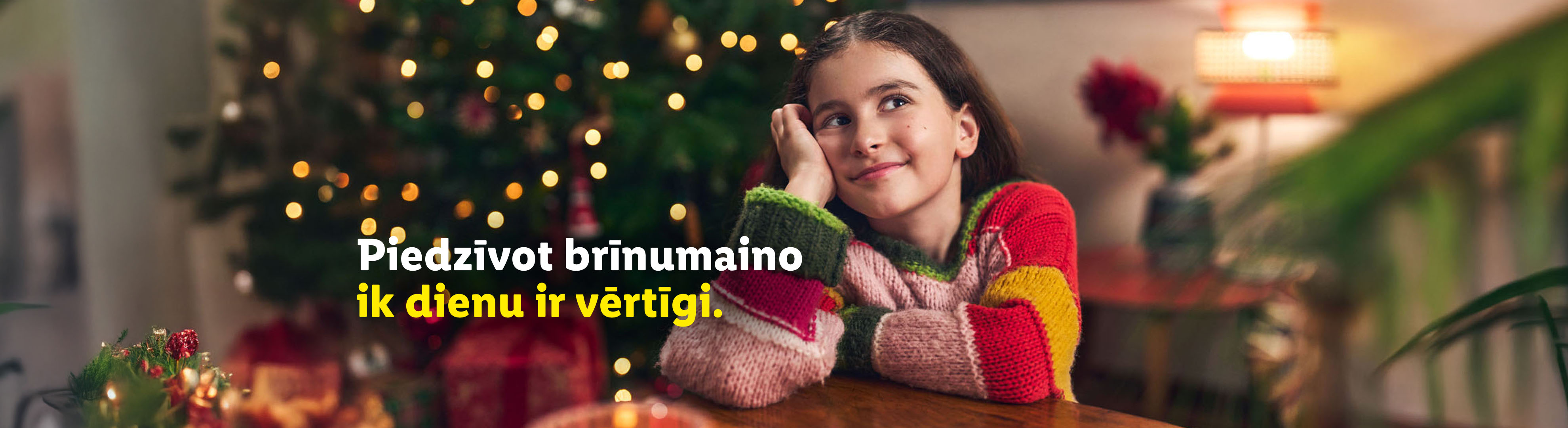 Meitene džemperī sēž pie galda, fonā eglīte ar virtenēm. Teksts: 'Piedzīvot ik dienu ir vērtīgi.'