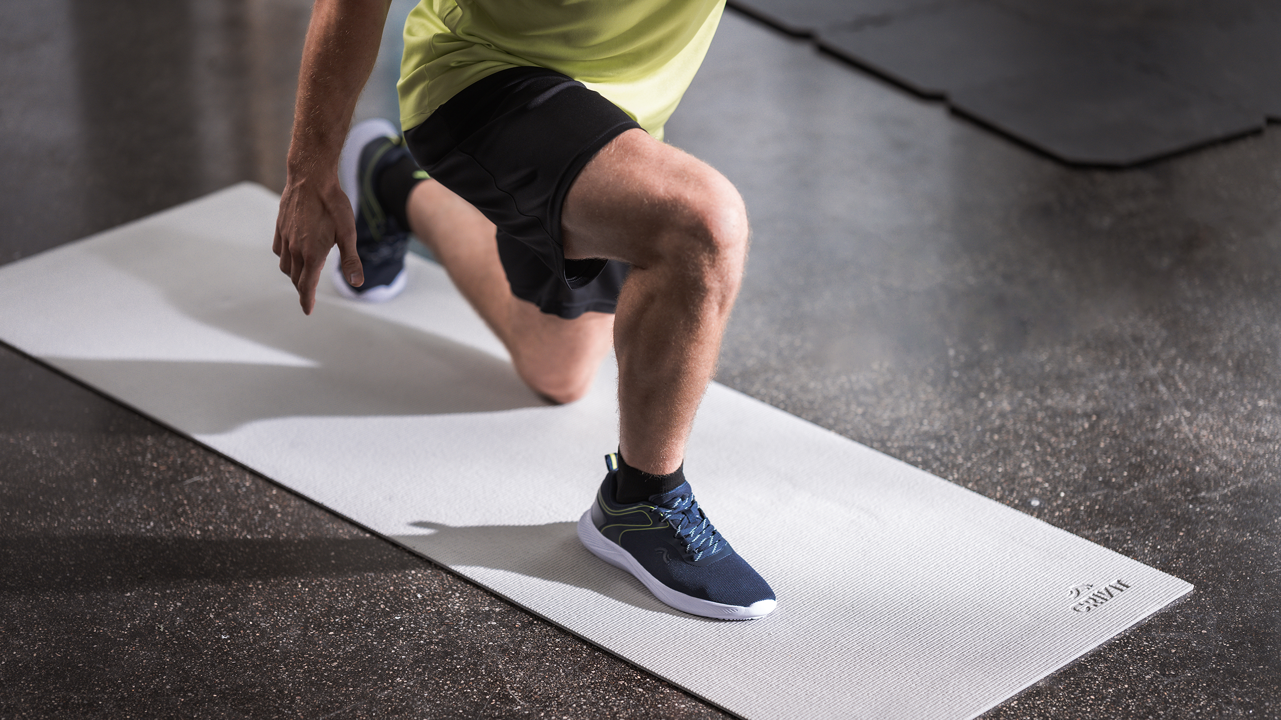 Мужчина в спортивной одежде делает выпады на светло-сером коврике для упражнений.