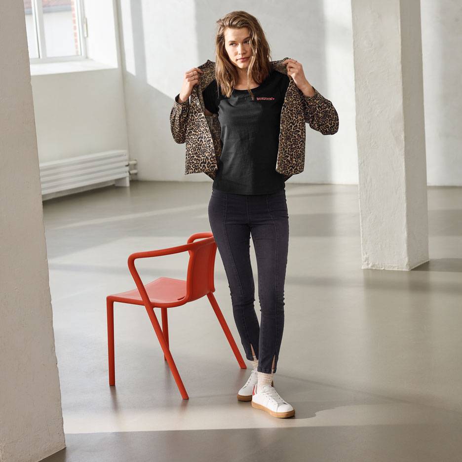 Sieviete melnā t-kreklā ar uzrakstu, džinsos un leoparda raksta jakā, stāv blakus sarkanajam krēslam.