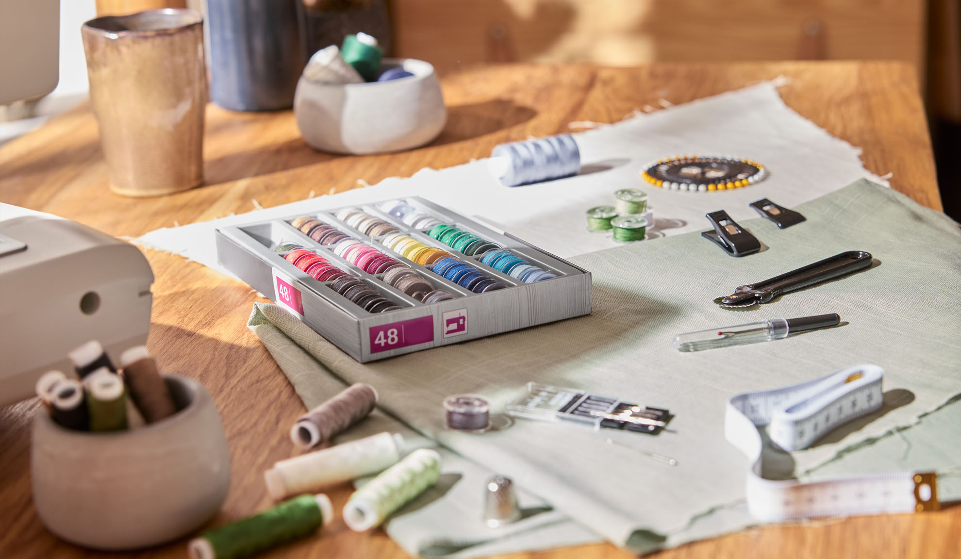 Šūšanas komplekts ar krāsainu diegu kasti, šujmašīnu un dažādiem instrumentiem uz koka galda.