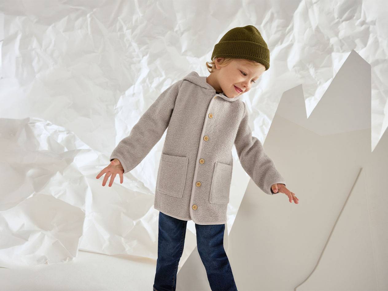 Мальчик в серой шерстяной куртке и зеленой шапке, улыбающийся на фоне мятой бумаги.