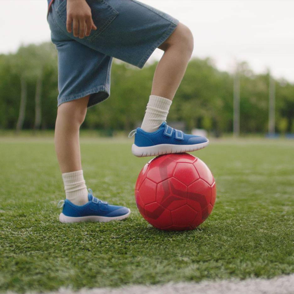 Bērns šortos un zilos sporta apavos, ar vienu kāju uz sarkanas futbola bumbas.