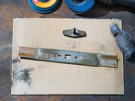 Sarūsējis zāles pļāvēja asmens uz kartona ar abrazīviem diskiem un tērauda vilnu.