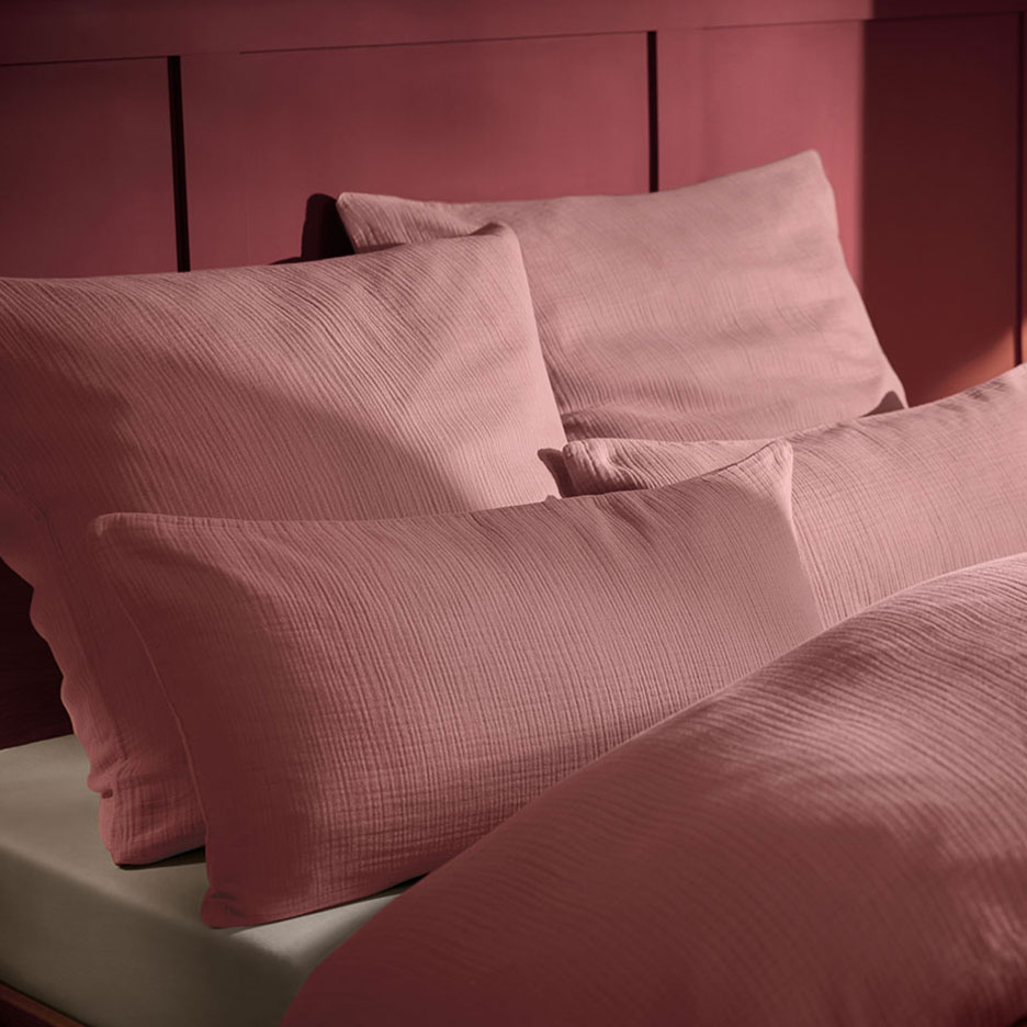 Розовая подушка и муслиновое постельное белье на кровати с деревянным изголовьем.