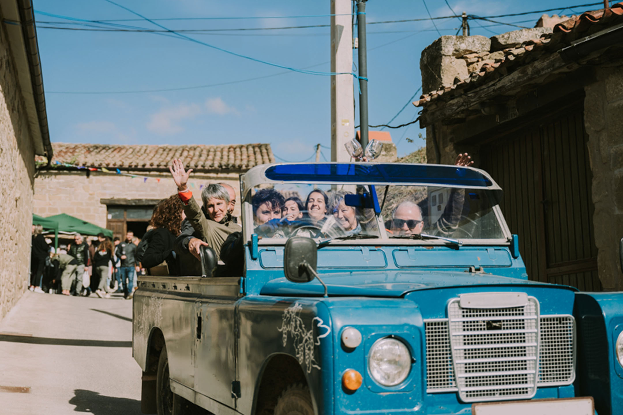 Menschen in einem blauen Oldtimer-Geländewagen fahren durch eine Dorfstraße.