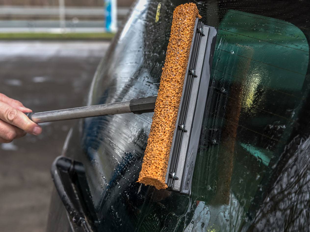 Logu tīrītājs ar garu rokturi tīra automašīnas aizmugurējo stiklu.