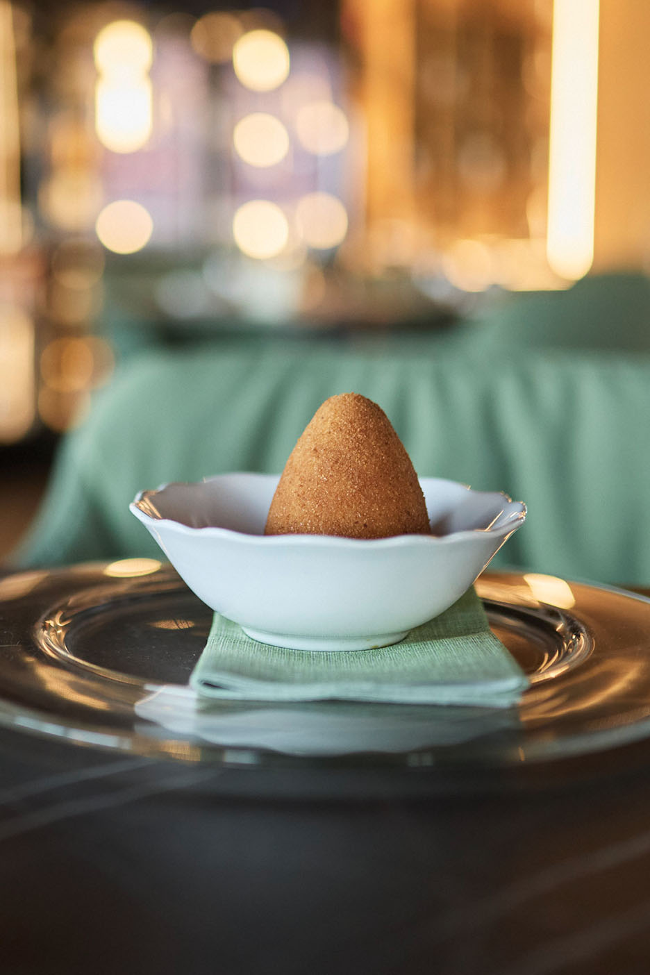 Zeltīts arancini baltā bļodā uz zaļas salvetes un stikla šķīvja.
