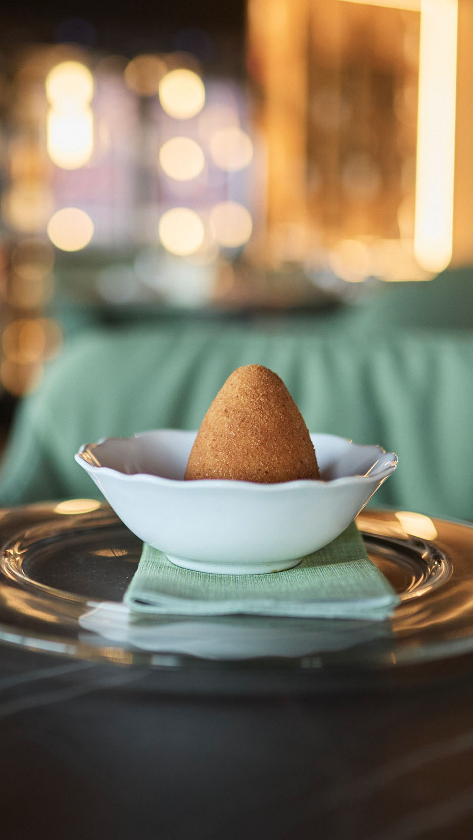 Zelta arancini baltā bļodā uz zaļas salvetes un stikla šķīvja.