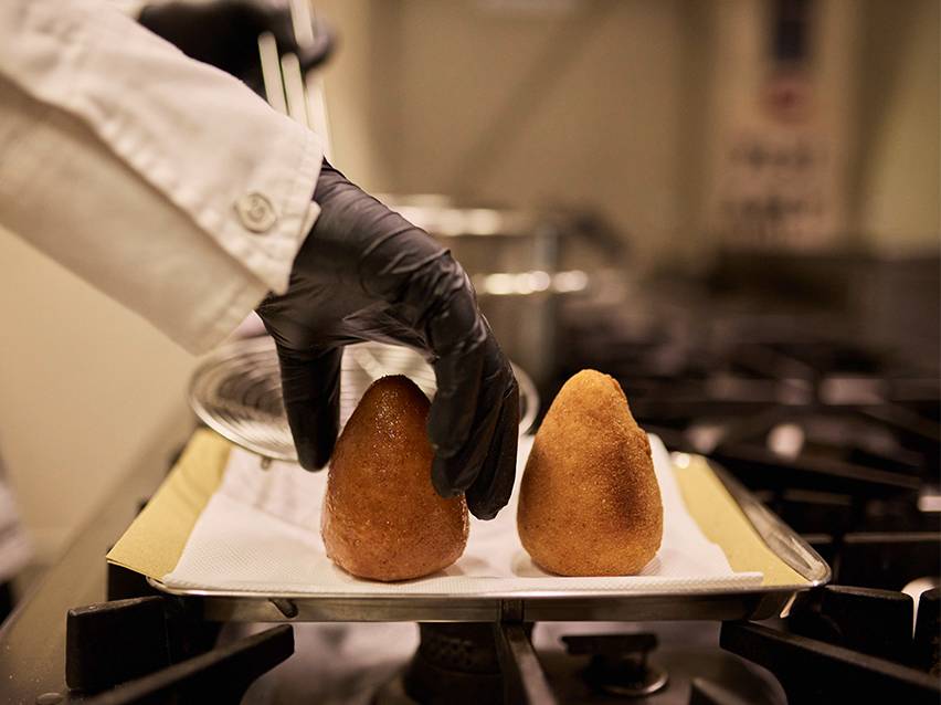 Pavārs melnos cimdos liek ceptus arancini uz papīra dvieļu paplātes.