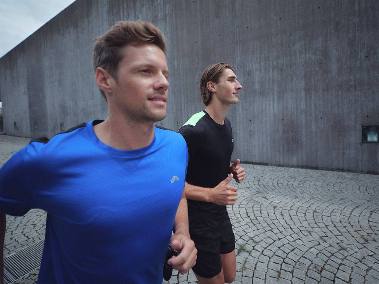 Двое мужчин бегут в спортивных футболках и шортах, один в синей, другой в черной.