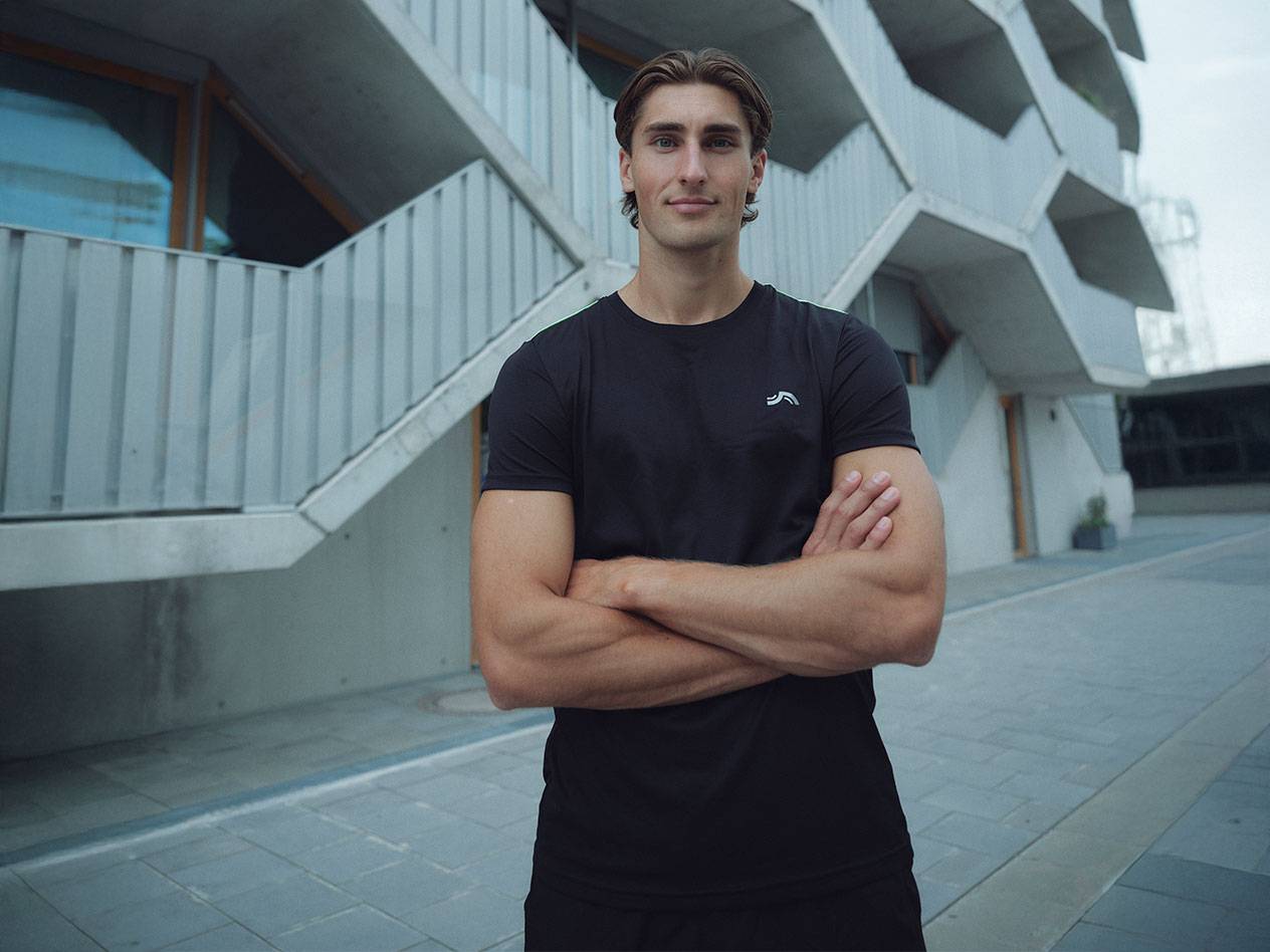 Мужчина в черной футболке с логотипом и черных шортах, стоит на улице, скрестив руки.