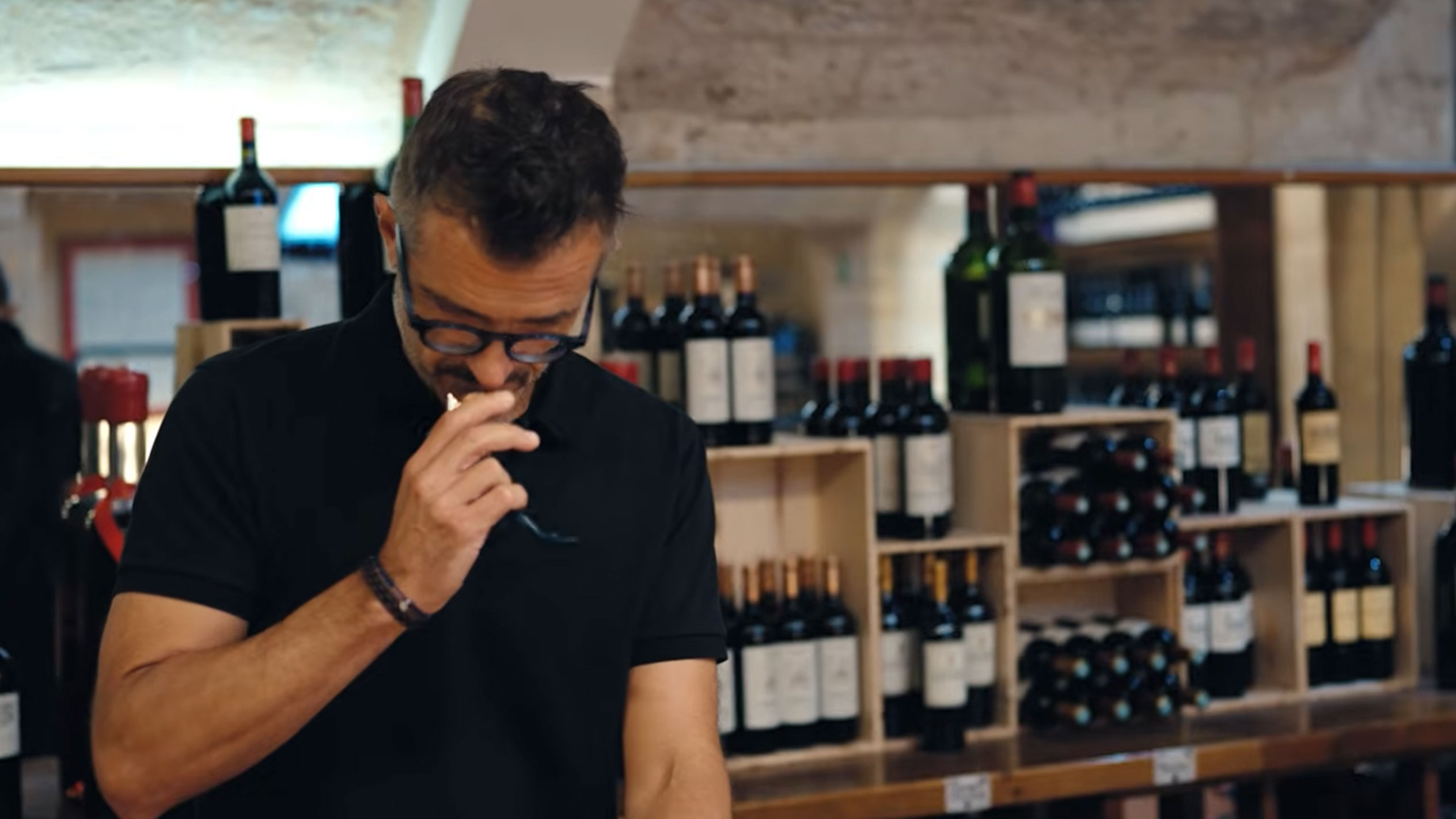 Mann in einem Weinkeller verkostet Wein, umgeben von Flaschen auf Holzregalen.