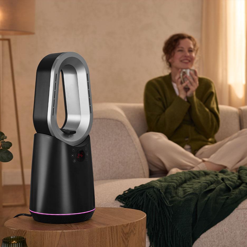 Bezspārnu sildītājs-ventilators uz galdiņa mājīgā viesistabā ar sievieti uz dīvāna.