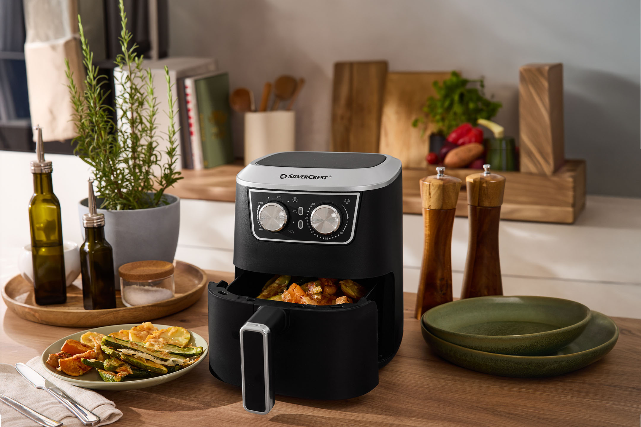 Черная аэрофритюрница с овощами, специями и посудой на деревянной столешнице.