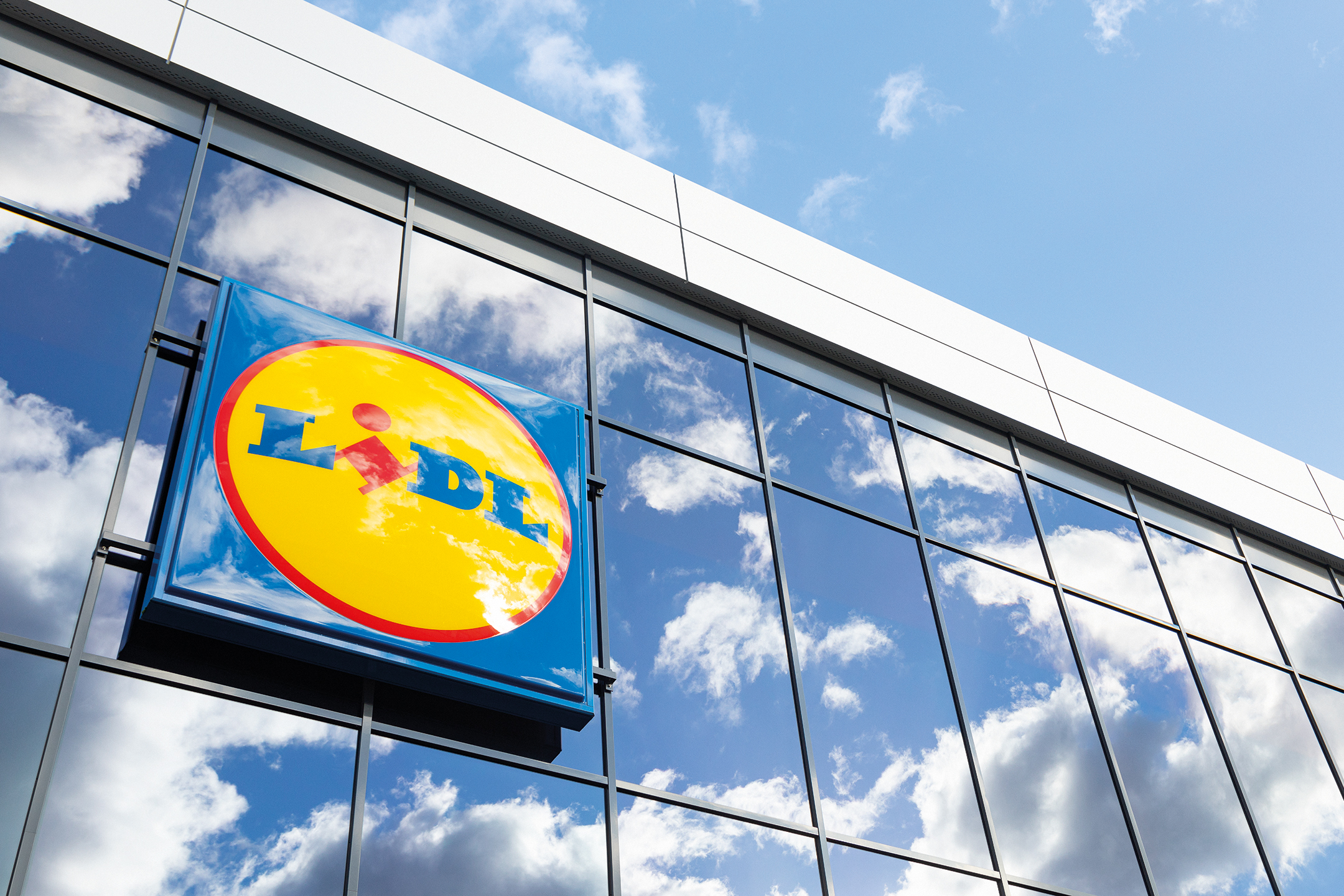 Lidl zīme uz stikla fasādes ar debesu un mākoņu atspulgu.