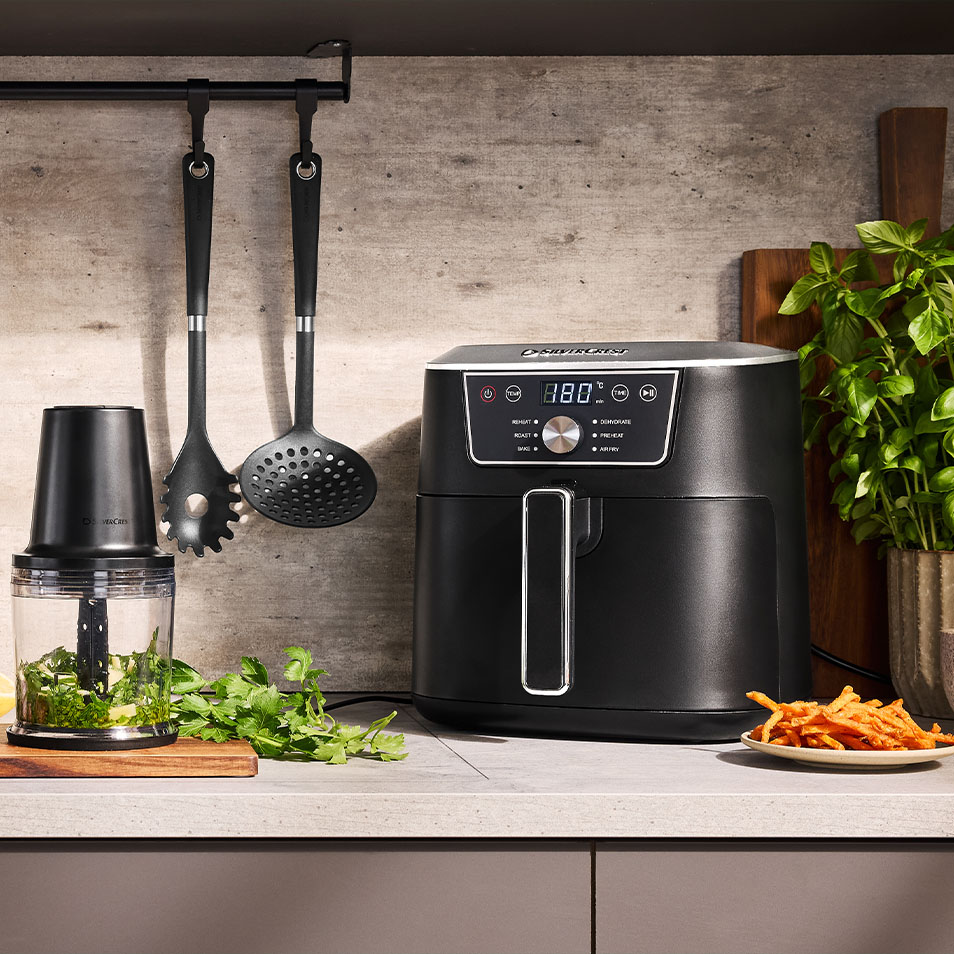 Аэрофритюрница SilverCrest и измельчитель продуктов на кухонной стойке со свежей зеленью и картофелем фри из сладкого картофеля.