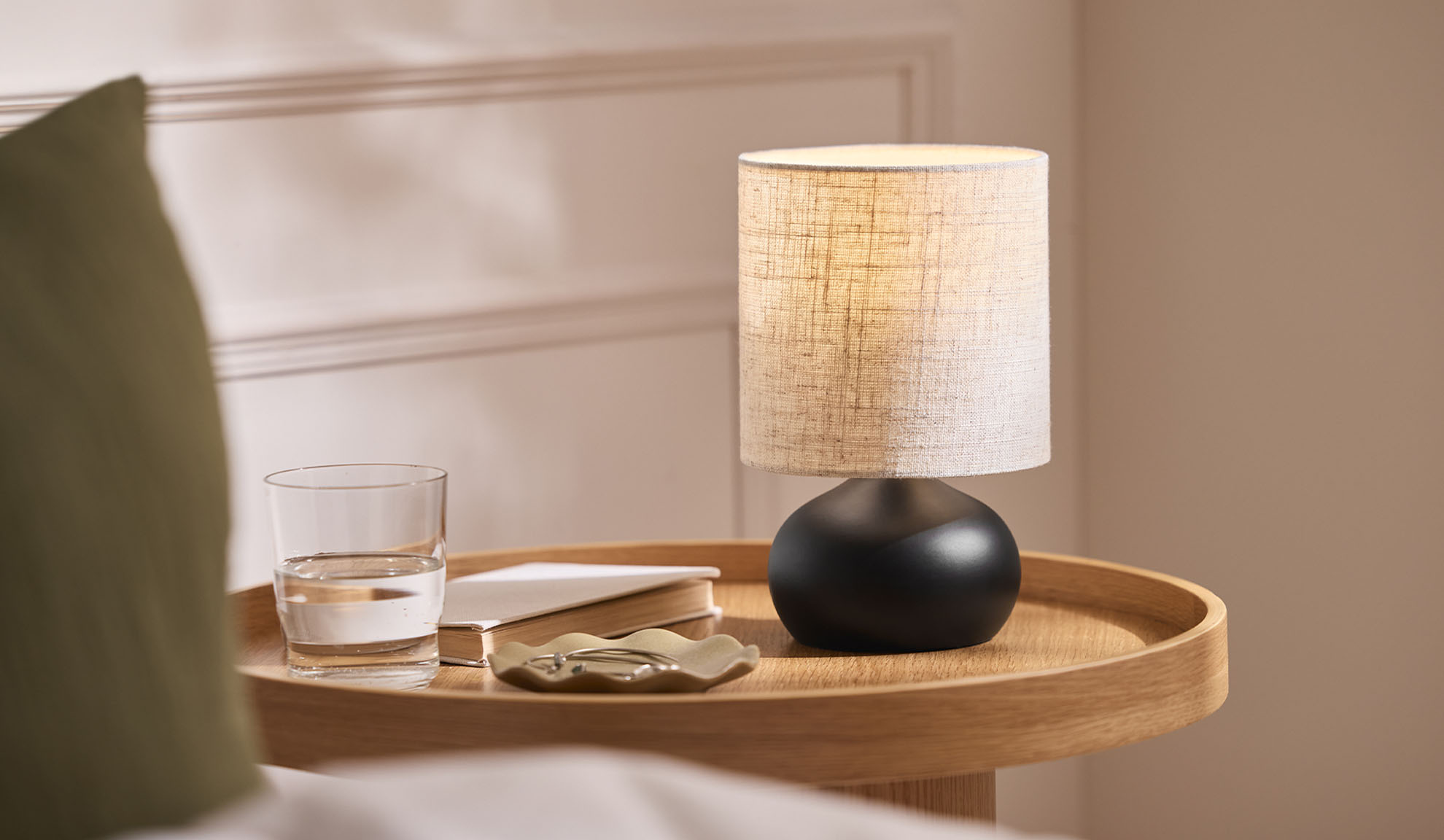 Naktsskapīša lampa, glāze, grāmata un rotaslietu paplāte uz koka paliktņa pie gultas.
