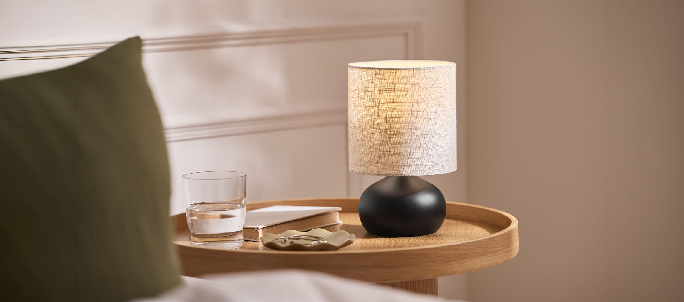 Gultas lampa ar auduma abažūru, glāze ūdens un grāmata uz koka paplātes.