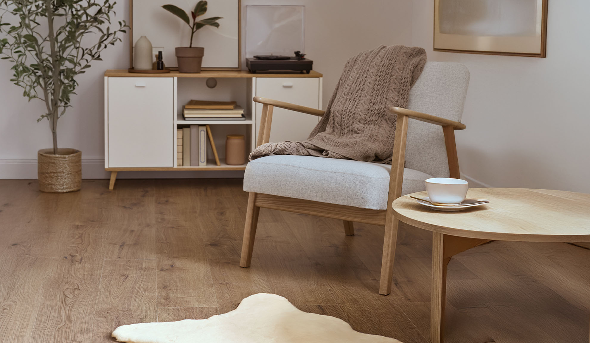 Mājīgs atpūtas krēsls ar pledu, koka kafijas galdiņu un uzglabāšanas skapi gaišā viesistabā.