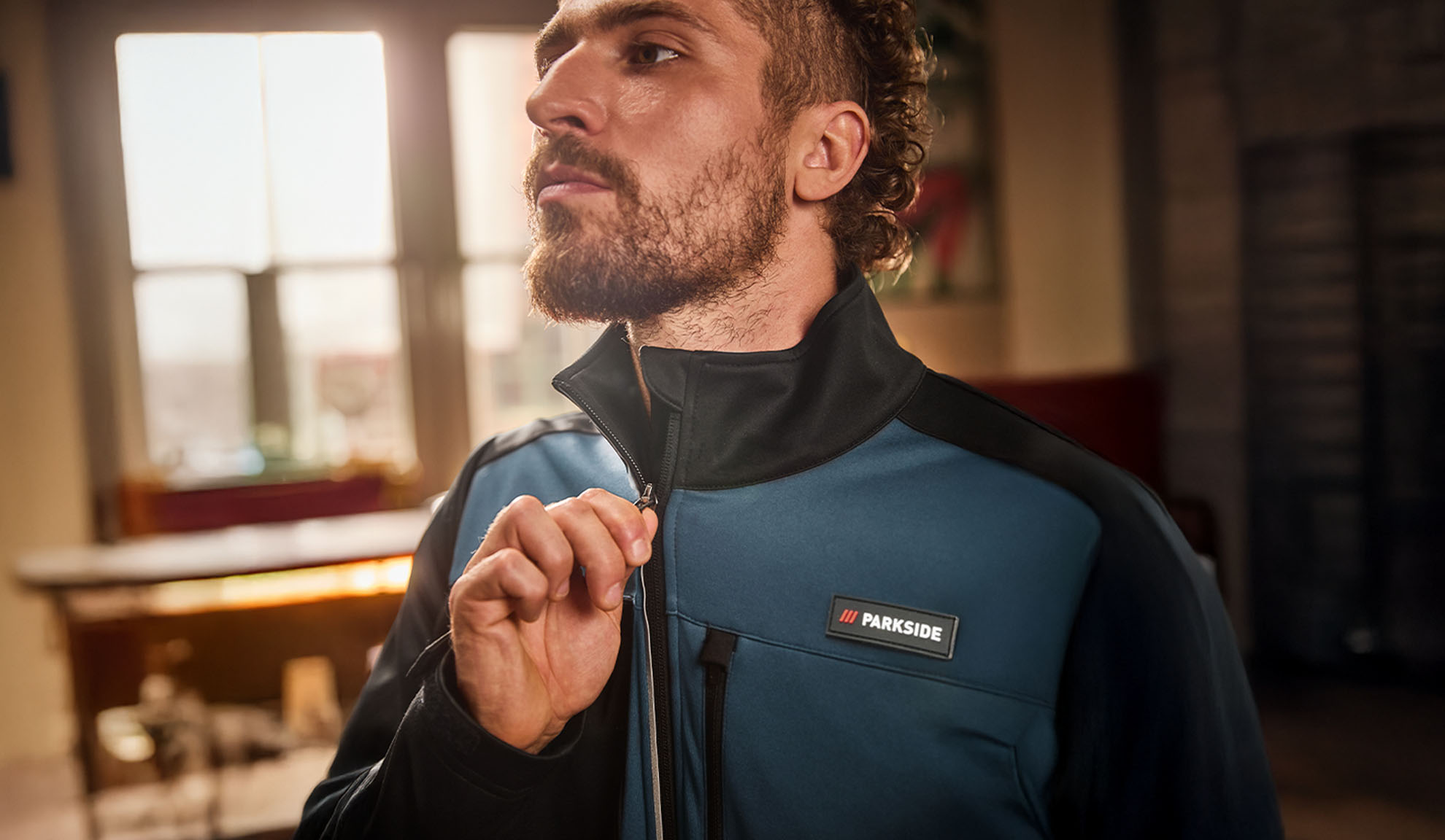 Vīrietis zilā un melnā Parkside darba jakā, aizver rāvējslēdzēju.