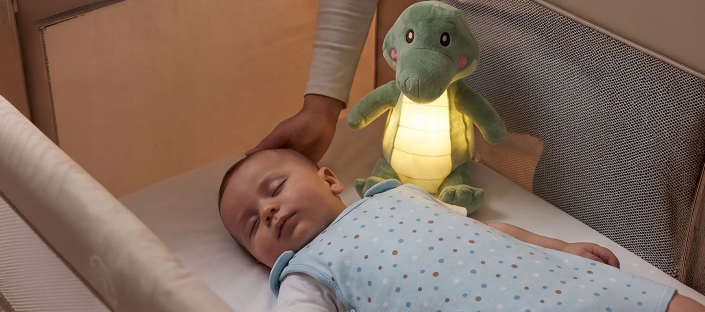 Mazulis guļ gultiņā ar mīkstu rotaļlietu-naktslampu, pieaugušais glāsta bērna galvu.
