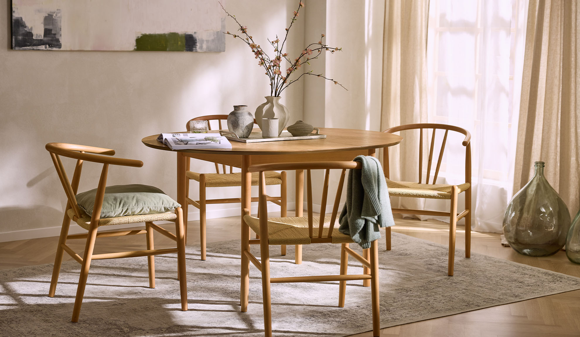 Gaiša ēdamistaba ar apaļu koka galdu, koka krēsliem ar virvju sēdekļiem un ziedu dekorācijām.