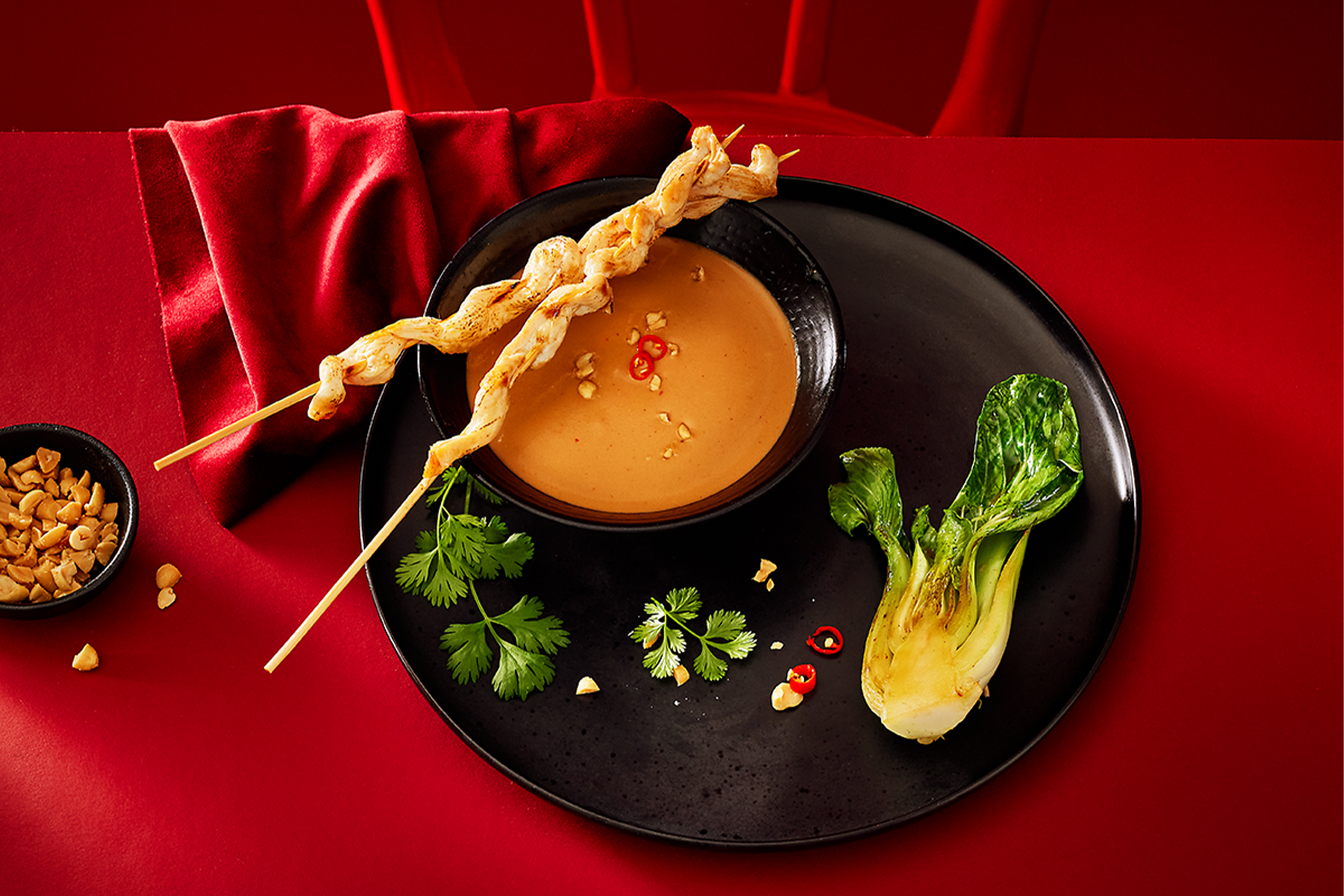 Vistas satay iesmiņi ar zemesriekstu mērci, pakčoju un koriandru uz sarkana fona.