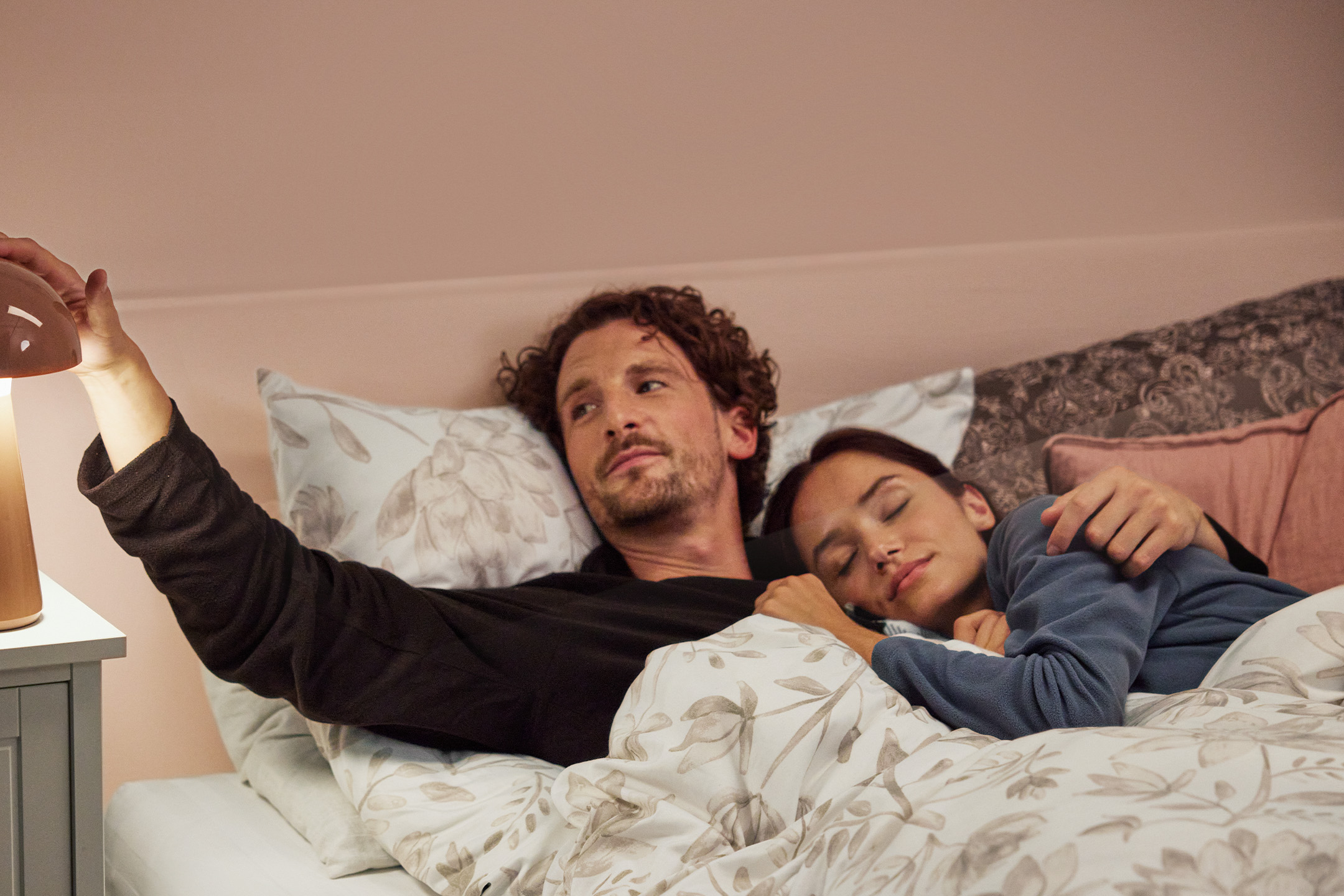 Pāris gultā ar smilškrāsas gultasveļu un naktsgaldiņu ar lampu