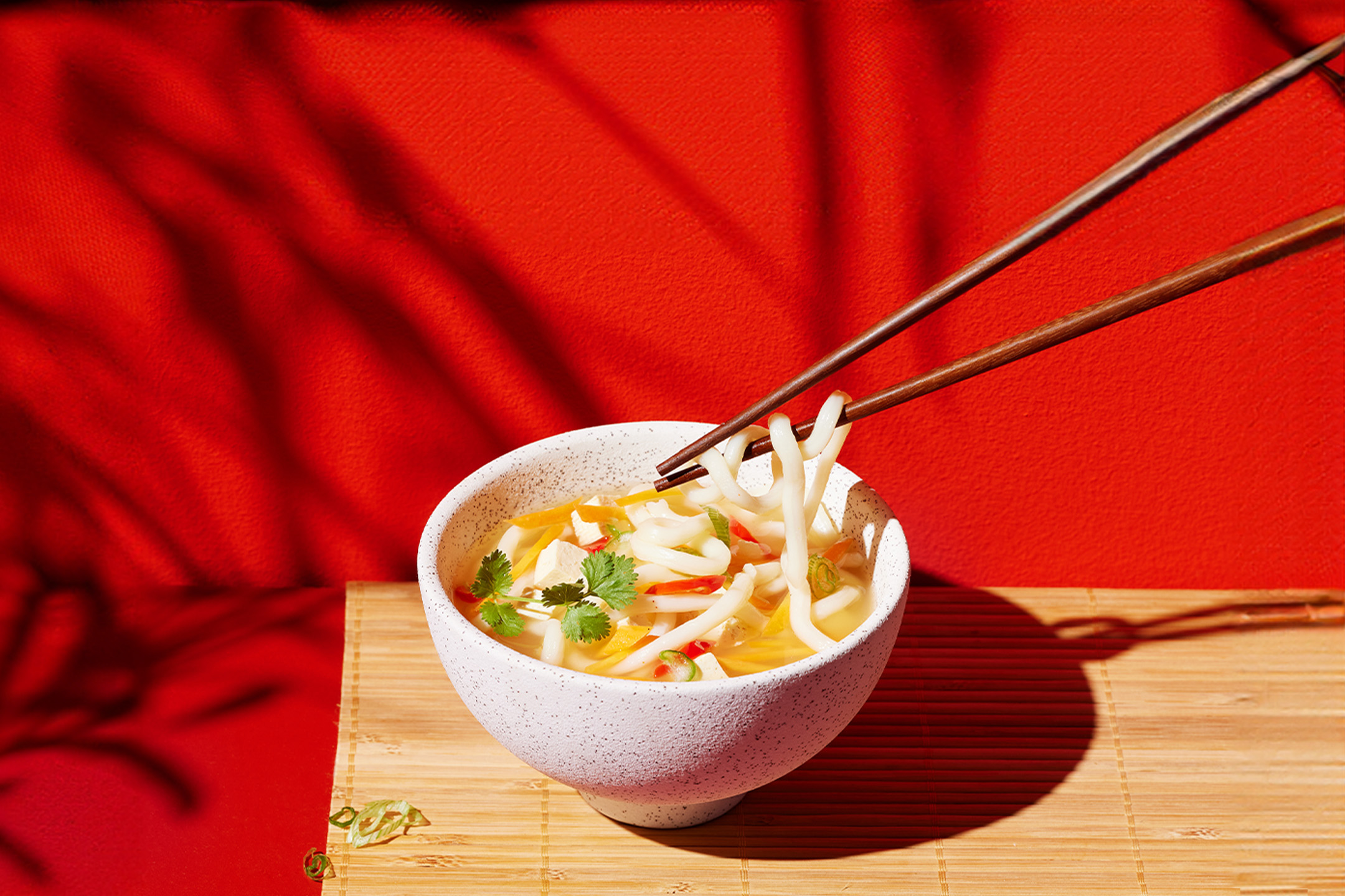 Bļoda ar udona nūdelēm, tofu, dārzeņiem un irbulīšiem uz bambusa paklājiņa.