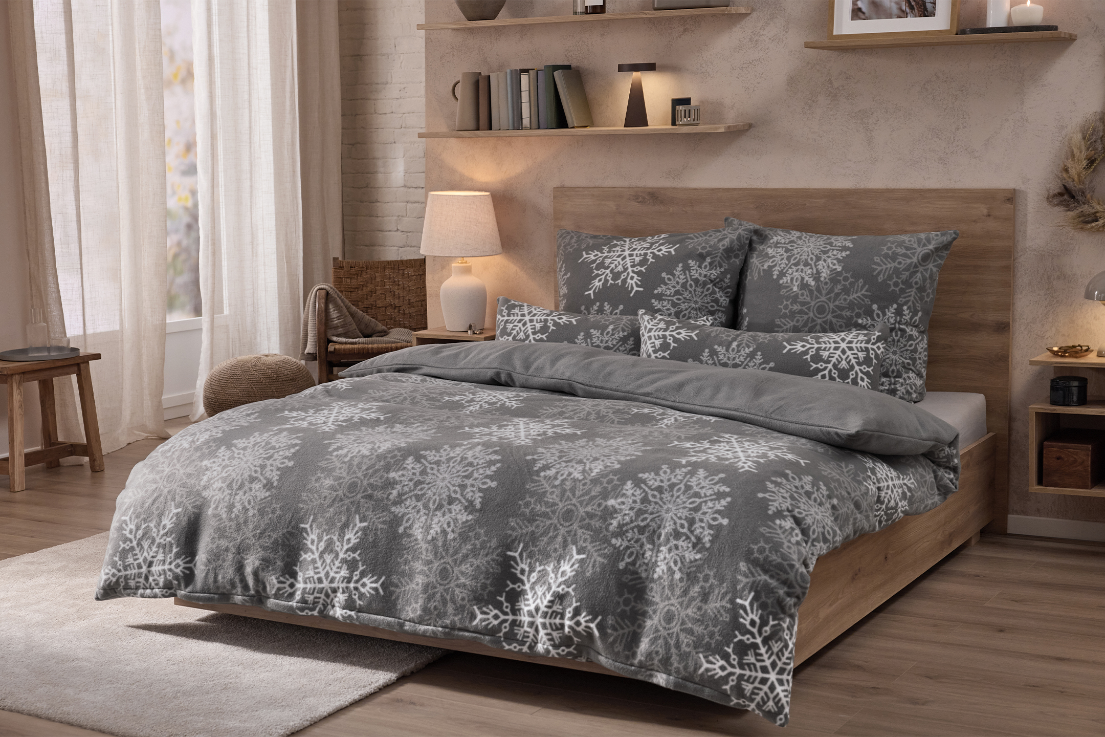 Guļamistaba ar gultu, pelēku gultas veļu ar sniegpārslu rakstu, koka mēbelēm un plauktiem.