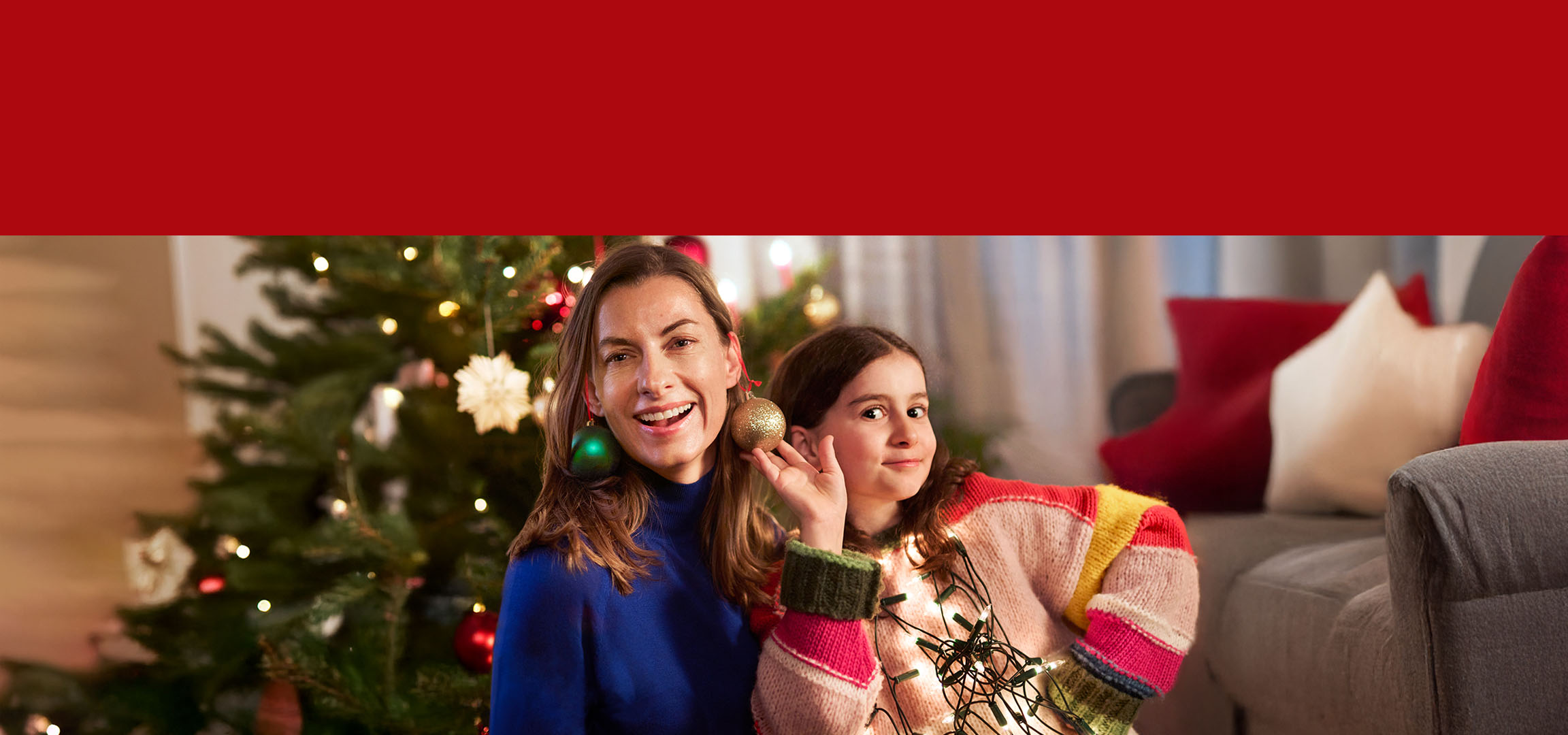 Sarkana Ziemassvētku bumba un sieviete ar meiteni pie eglītes.