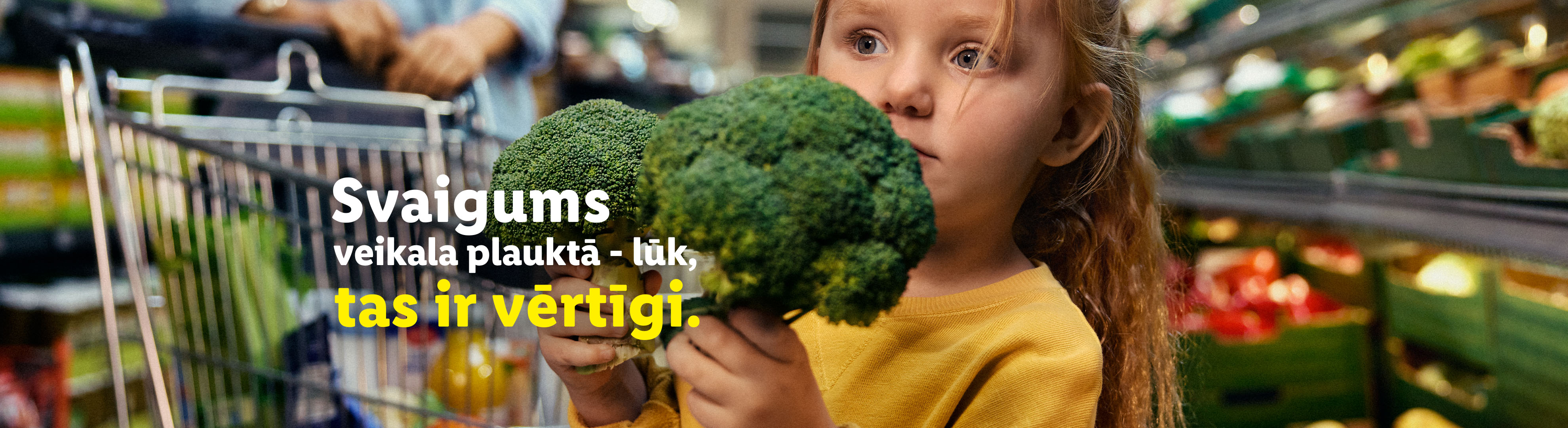 Meitene veikalā tur brokoļus, fonā iepirkumu ratiņi un dārzeņi. Teksts: 'Svaigums veikala plauktā – lūk, tas ir vērtīgi.'