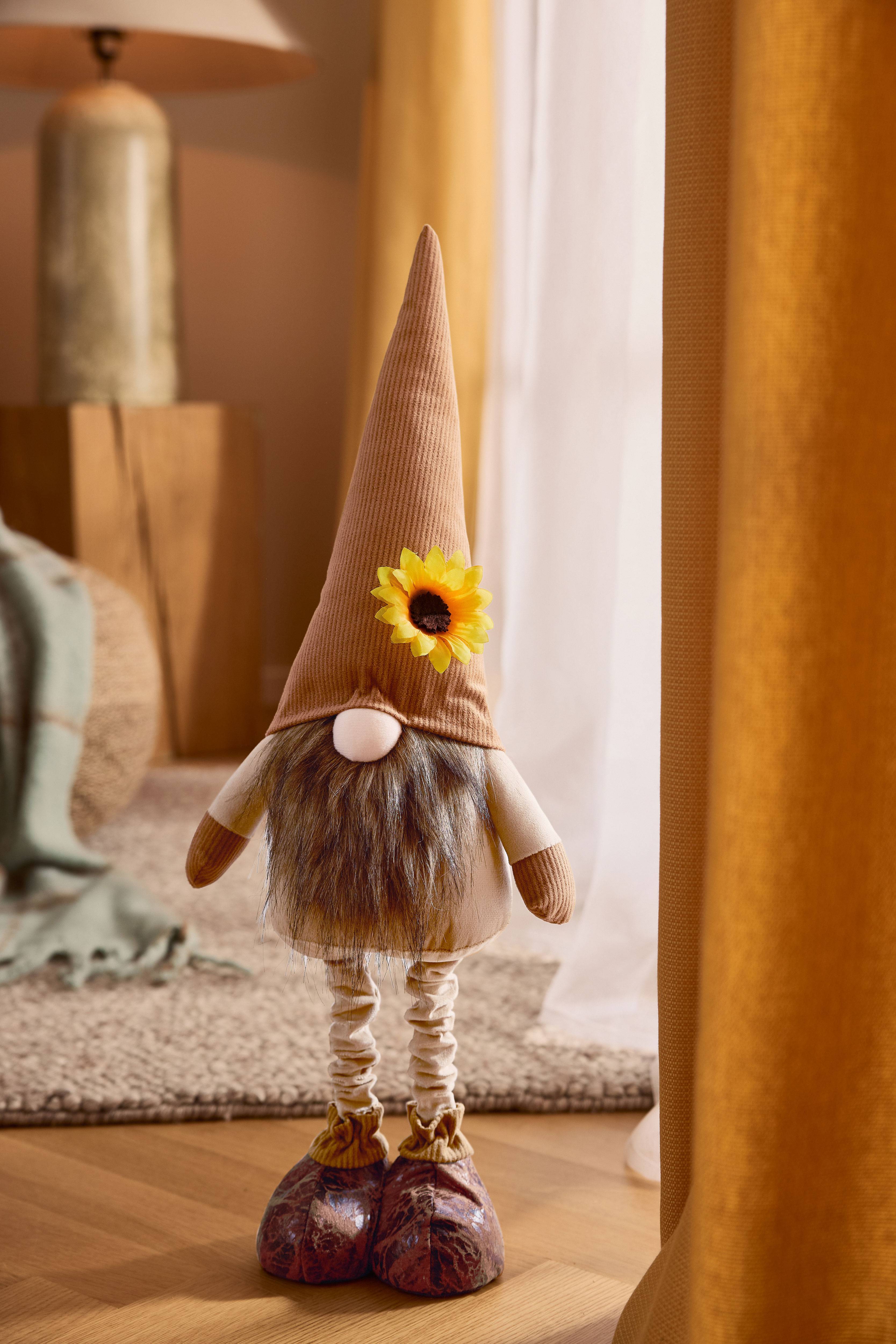 Плюшевый гном с коричневой остроконечной шляпой, бородой и подсолнухом, в уютной обстановке.