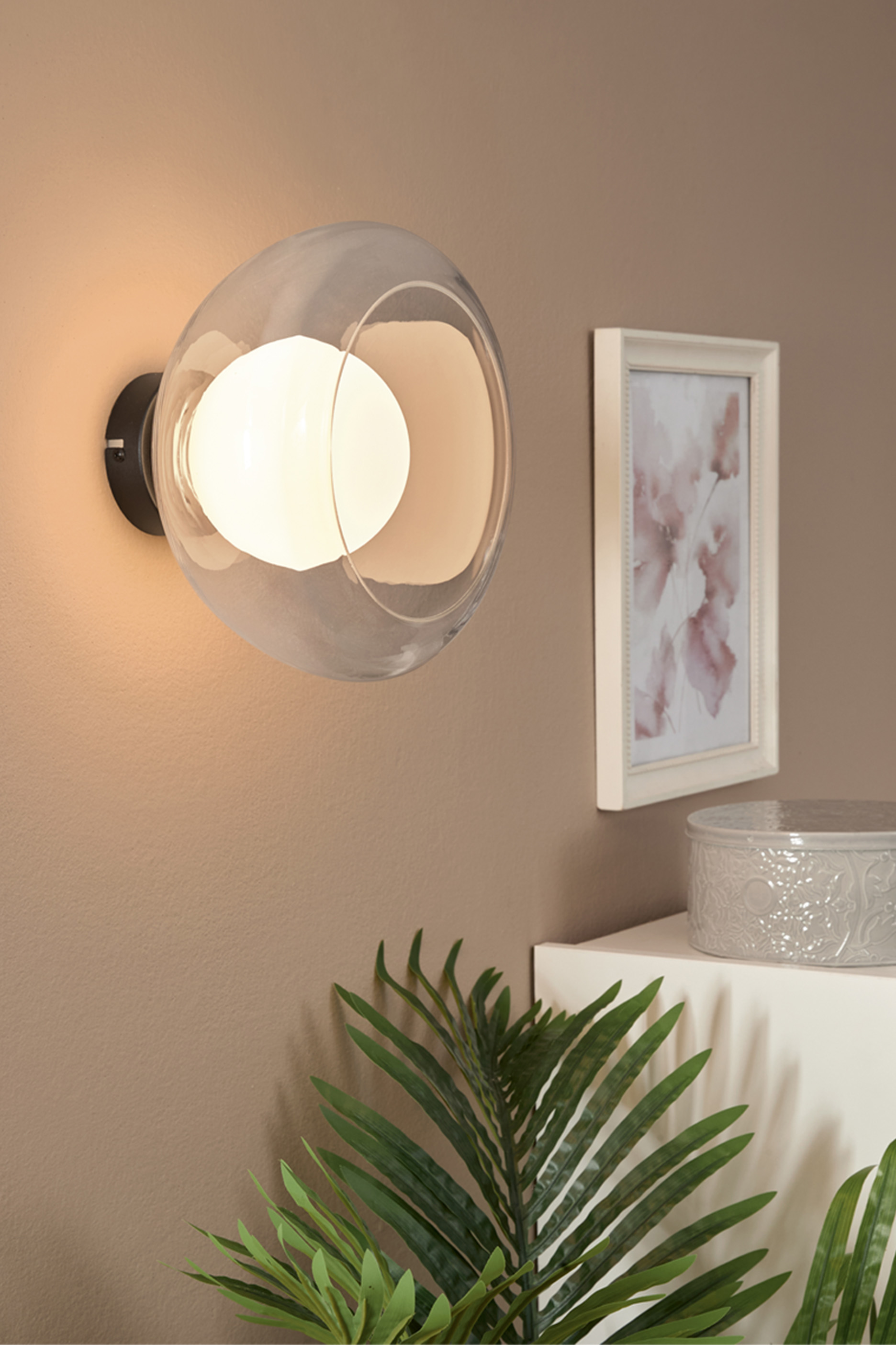 Sienas lampa ar caurspīdīgu abažūru, glezna ar ziediem un dekoratīva kaste uz plaukta.