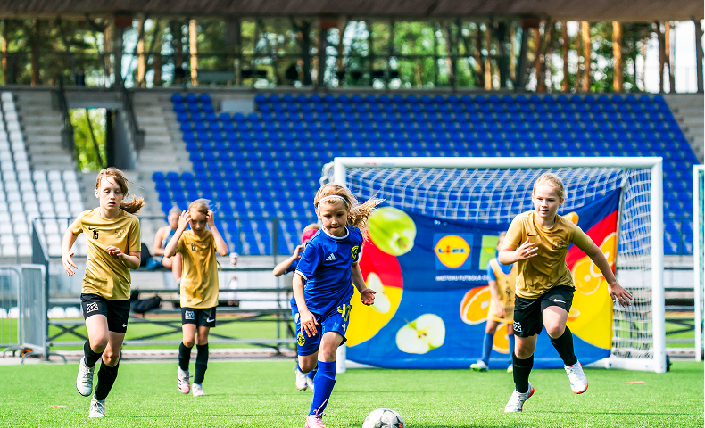 Bērni spēlē futbolu uz zaļa laukuma ar vārtiem un tribīnēm fonā.