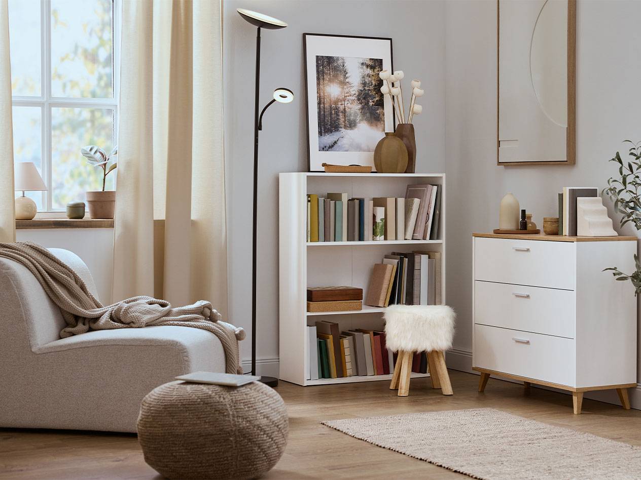 Уютная гостиная с креслом, книжным шкафом и комодом в нейтральных тонах.