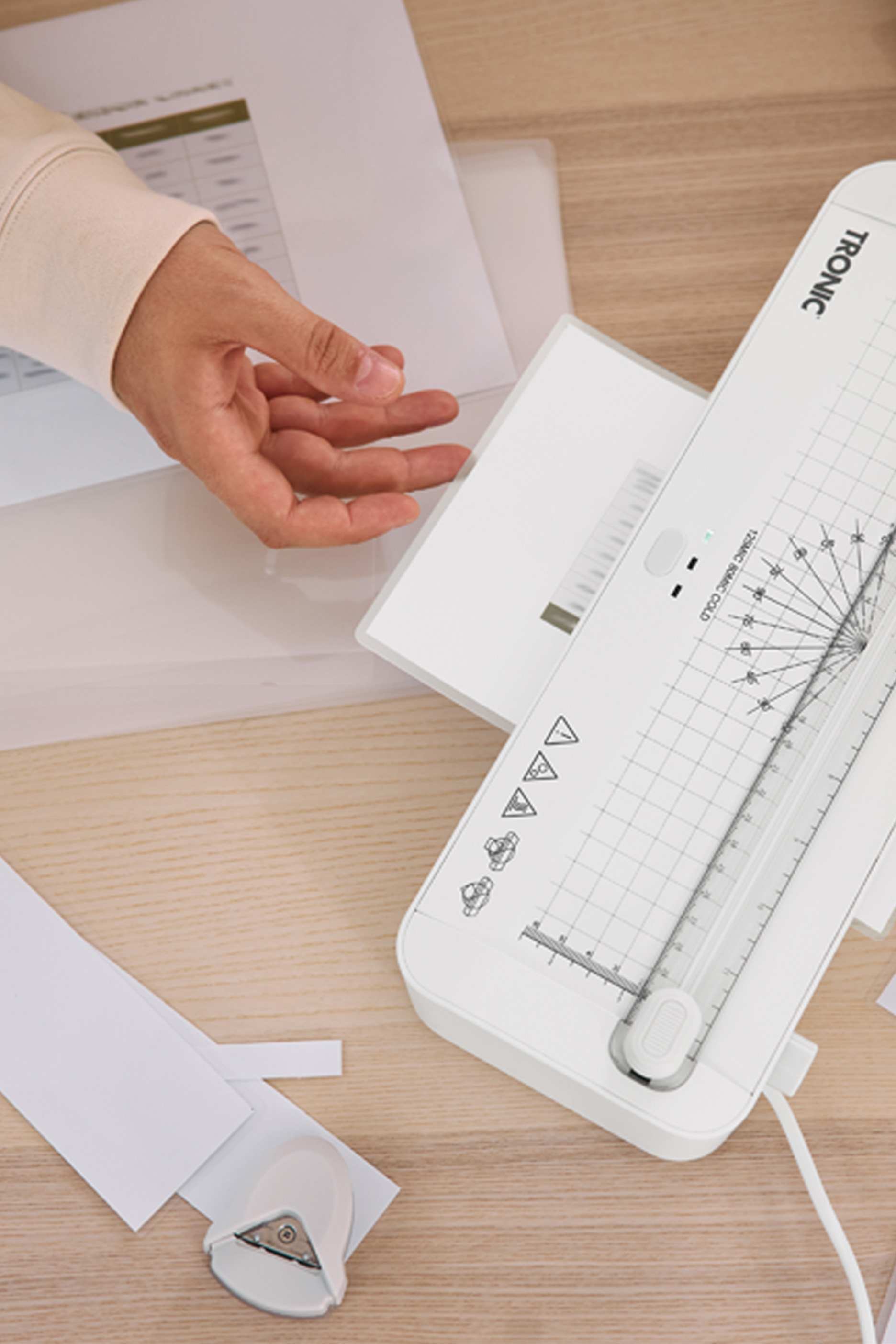 Cilvēka roka, kas izmanto Tronic laminatoru, blakus papīram un stūra caurumotājam.