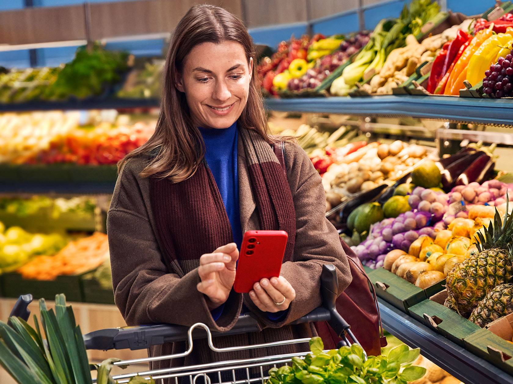Sieviete ar viedtālruni pārtikas veikalā, iepērkoties starp augļu un dārzeņu plauktiem.