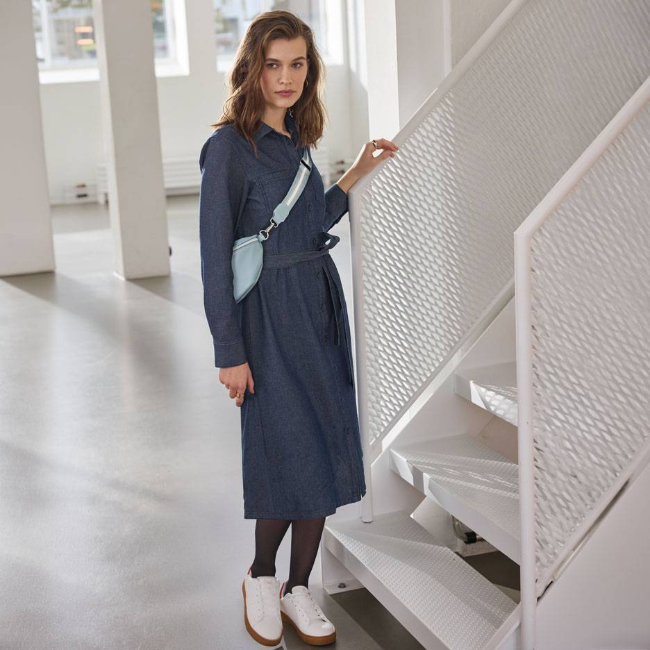 Sieviete džinsa krekla kleitā, melnās zeķbiksēs un baltos sporta apavos, ar gaiši zilu jostas somu.