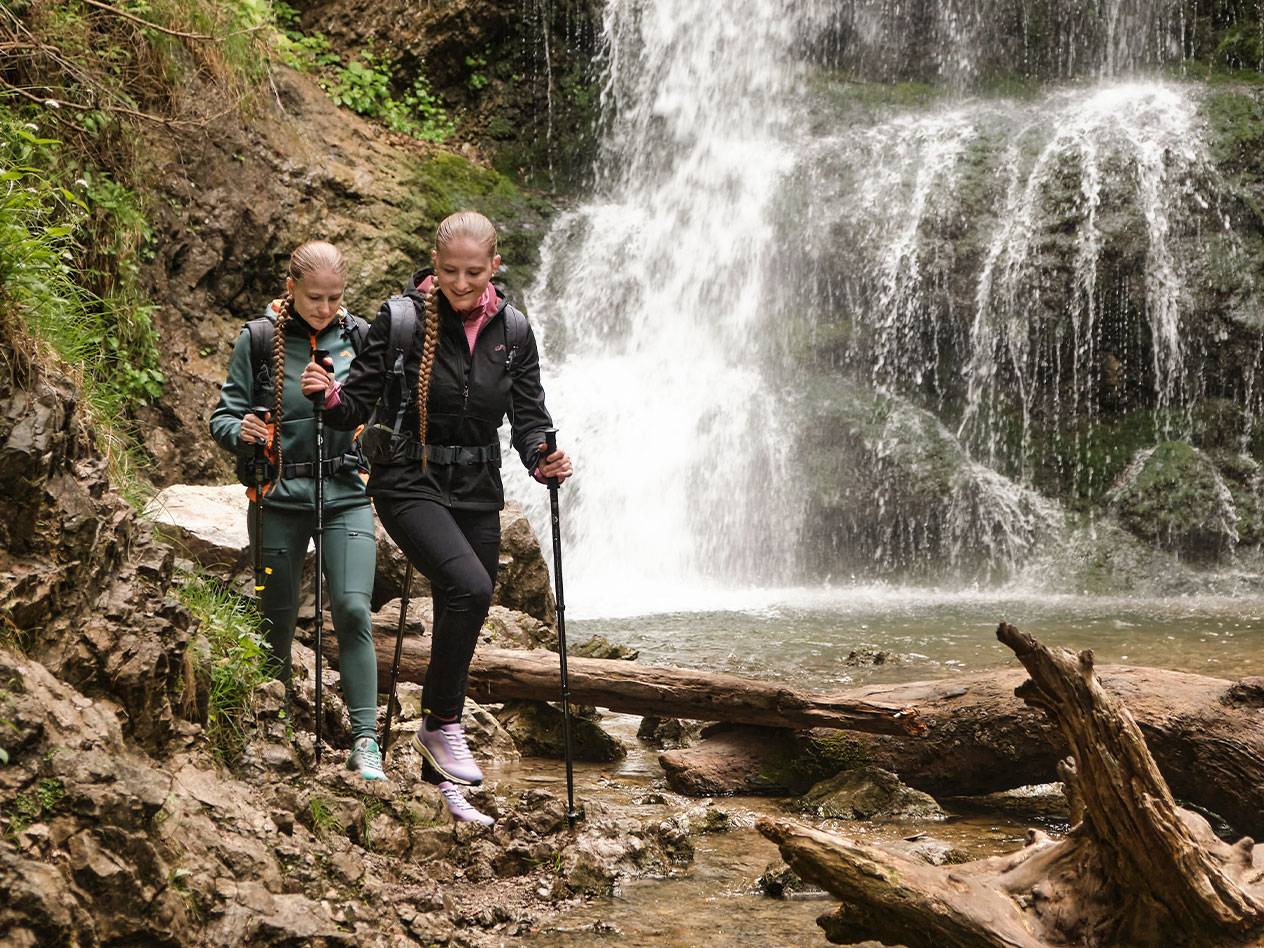 Divas sievietes ar trekinga nūjām iet pa akmeņainu taku pie ūdenskrituma.