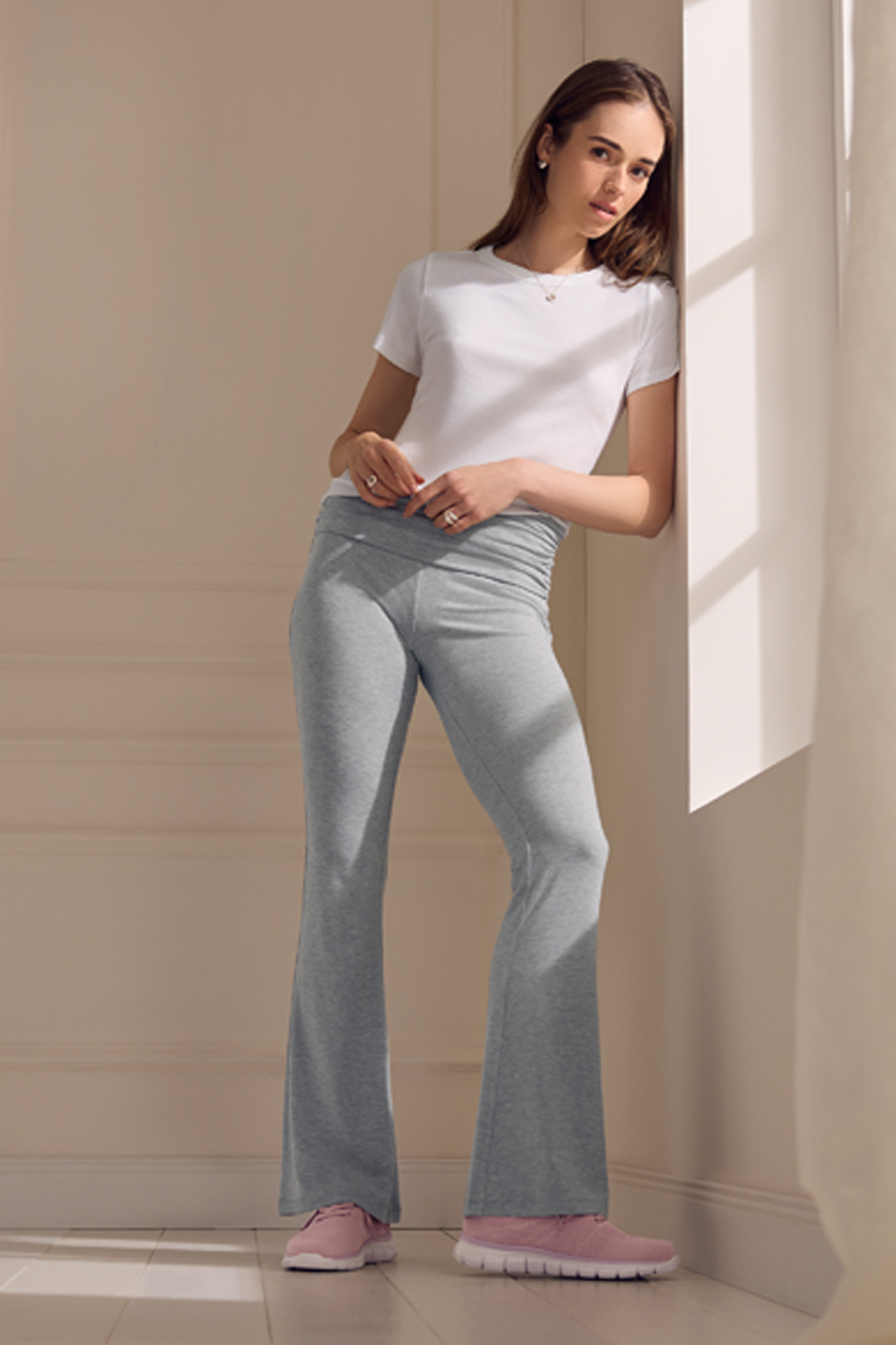 Sieviete baltā T-kreklā un pelēkās sporta biksēs, rozā kedās, atspiedusies pret sienu.